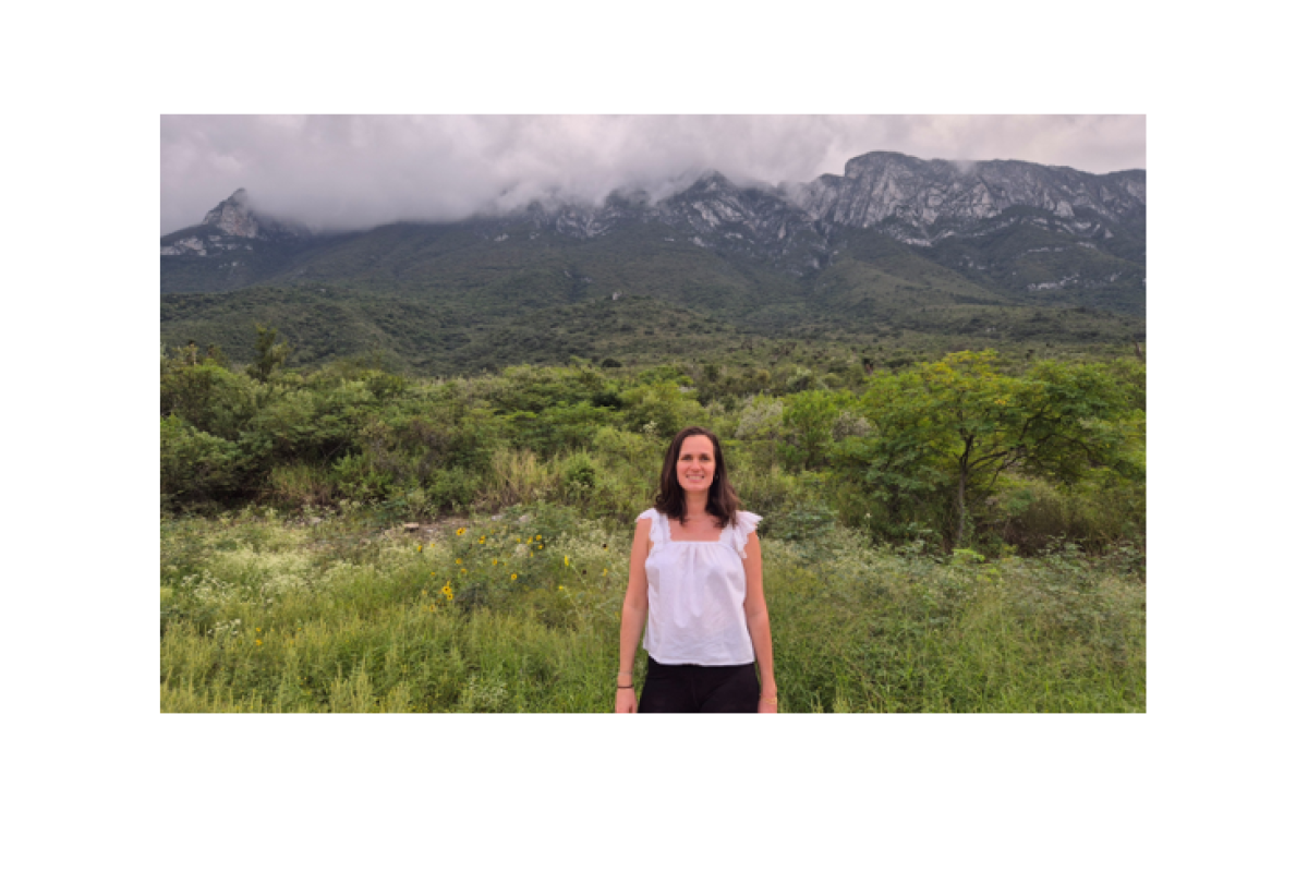 Eine Frau in einer weißen Bluse steht vor einer nebligen Gebirgskette und schaut lächelnd in die Kamera.