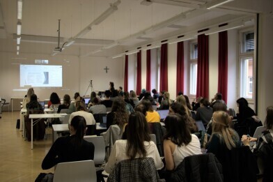 Viele Personen sitzen in einem Hörsaal und blicken auf die eingeblendete Präsentation